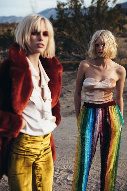 Two women in colorful fashion in a desert landscape. One wears an orange coat, the other a tube top and rainbow sequin pants.