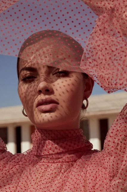 Close-up portrait of a young woman in a pink polka dot dress, with a matching sheer polka dot scarf draped over her head and shoulders.