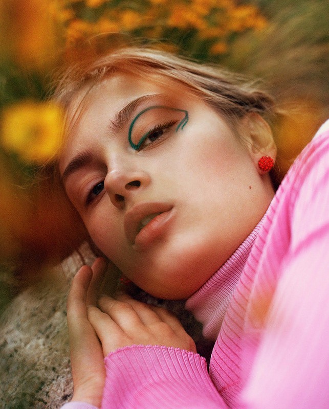 Close-up portrait of a fair-skinned young woman with green eyeliner, red earrings, and a pink top, lying among yellow flowers.