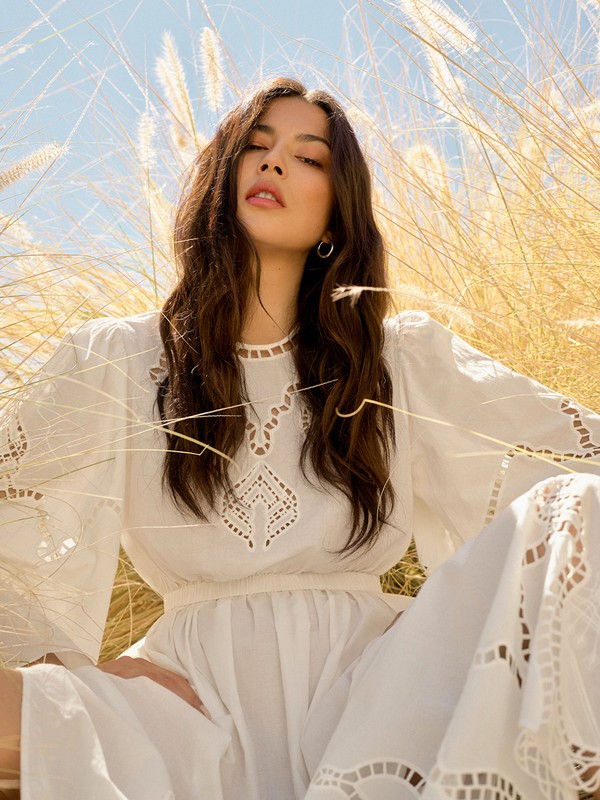 Woman with long dark hair wearing a white cutout dress framed by tall golden grass under a bright blue sky.