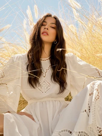 Woman with long dark hair wearing a white cutout dress framed by tall golden grass under a bright blue sky.