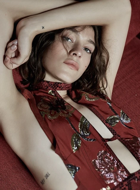 Brunette woman with floral-print red dress laying on red sofa with a neutral facial expression in an eye-level close-up studio shot.