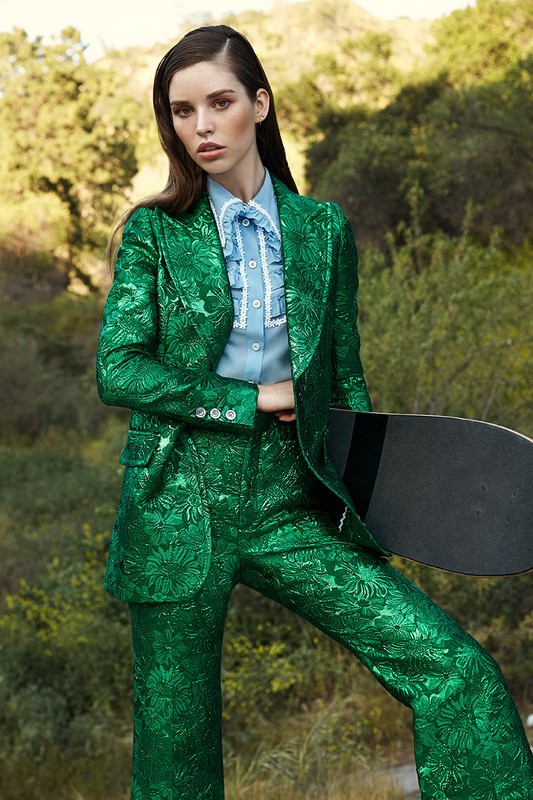 Model in a green floral suit and blue ruffled shirt holds a skateboard in an outdoor setting with trees and natural light.