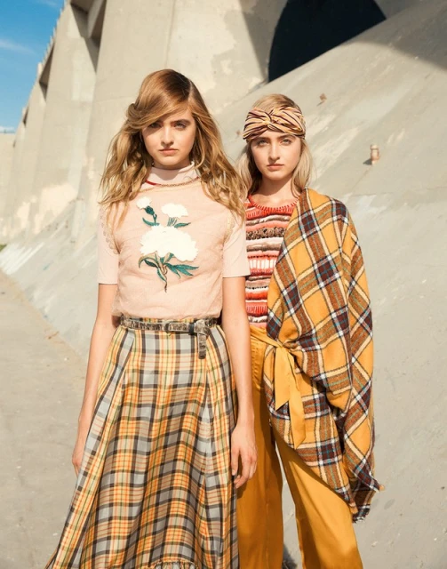 Two women in stylish vintage-inspired fashion stand in front of a concrete structure on a sunny day, showcasing retro-chic looks.