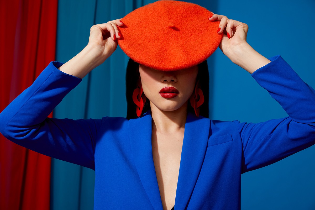 Woman in blue blazer and orange beret against a blue and red backdrop in a fashion-forward studio portrait.