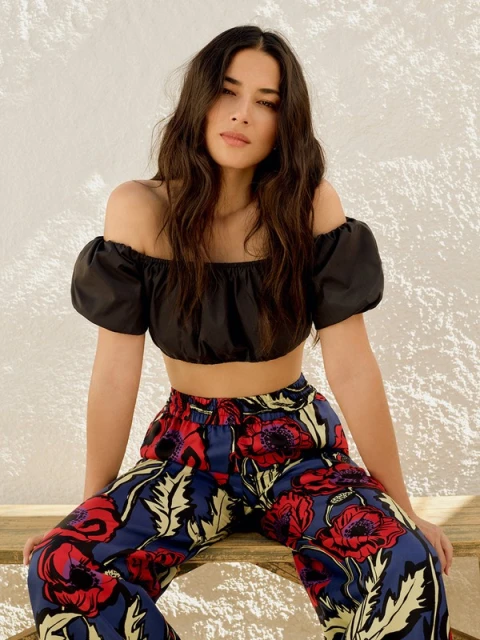 A young woman with long wavy hair poses while sitting; she wears a black off-shoulder crop top and floral print trousers against a light wall.
