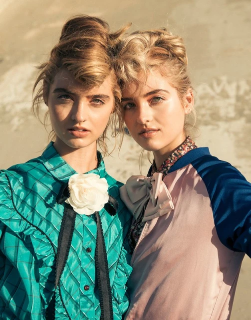 Two fair-skinned young women pose in satin tops against a blurred, textured background. Close up, fashion-editorial studio shot.