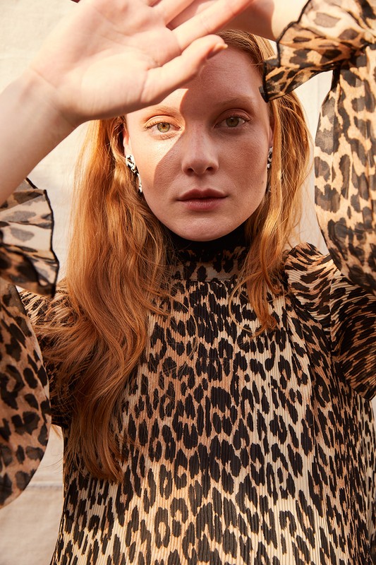 Close-up of a red-haired woman in a leopard-print blouse shielding her face from the sun, posing in an outdoor setting, fashion shoot.