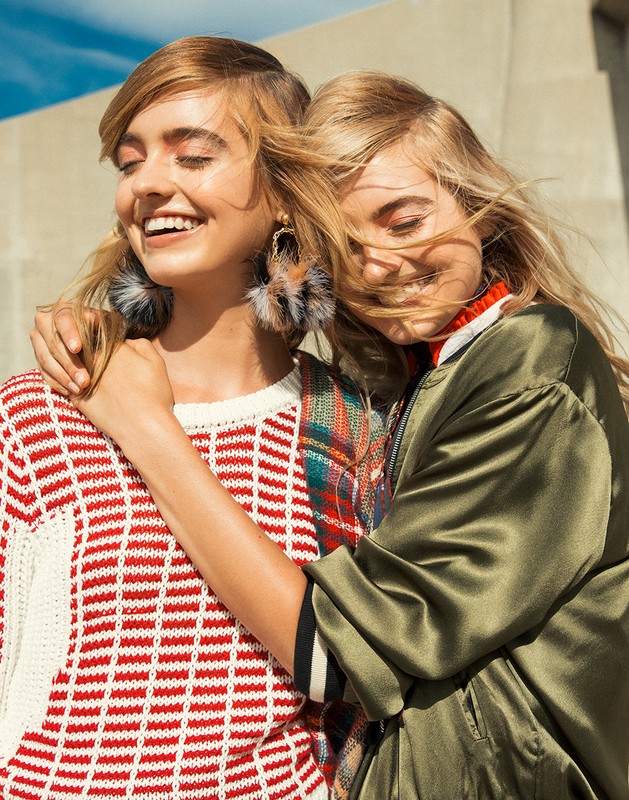 Two smiling women embrace outdoors, wearing fashionable sweaters and a jacket in natural light, creating a warm, friendly image.