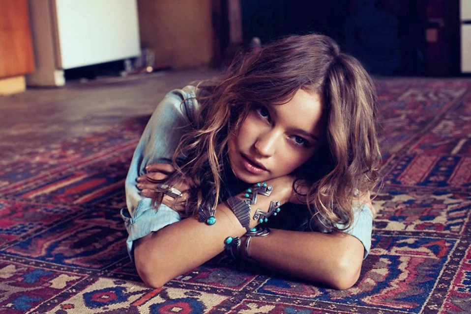Young woman with wavy brown hair, wearing a denim shirt and jewelry, lying on a patterned rug with wood floors in the background.