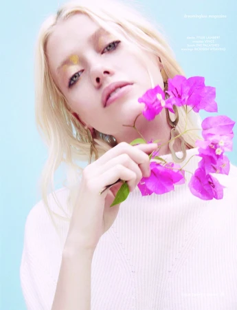 Close-up portrait of a blonde woman holding bright pink bougainvillea flowers, wearing a white textured sweater against a light blue background.