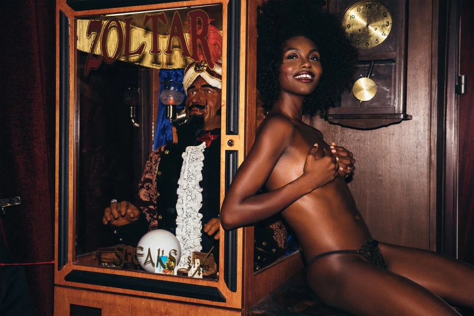 Young African American woman sitting near a Zoltar fortune teller machine in dimly lit room, smiling with hand covering breasts.