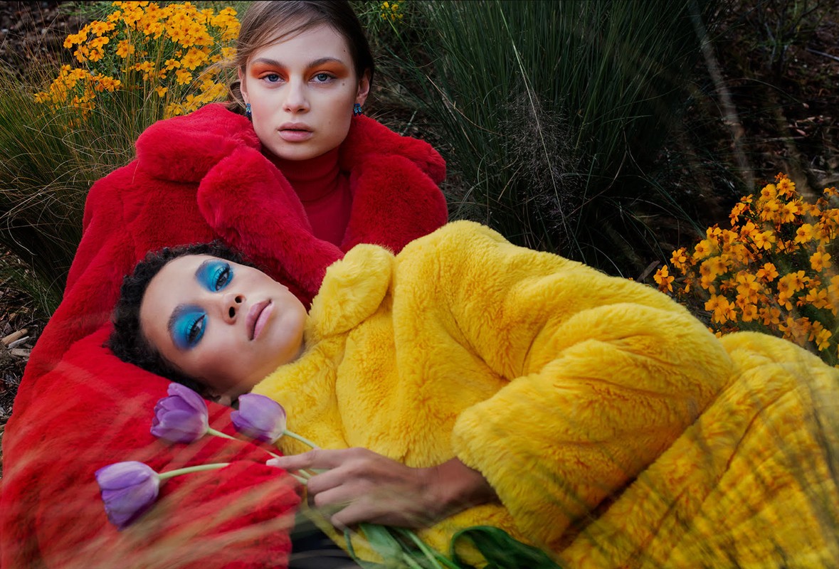 Two women in colorful faux fur coats pose outdoors surrounded by lush greenery and yellow flowers. Bright blue makeup accentuates their eyes.