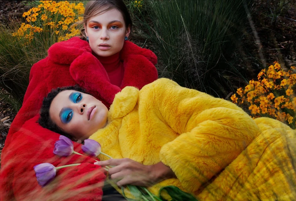 Two women in colorful faux fur coats pose outdoors surrounded by lush greenery and yellow flowers. Bright blue makeup accentuates their eyes.