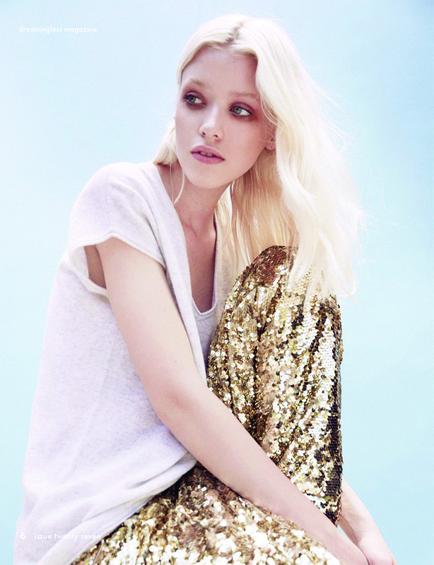 Young adult woman poses in a light-colored t-shirt and gold sequined pants against a soft blue background in a fashion studio shot.