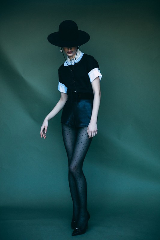 Model wearing a black wide-brimmed hat, black top, and polka dot tights against a green backdrop.
