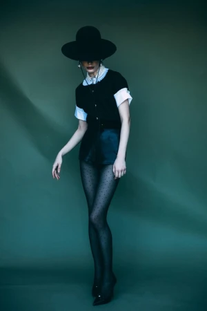 Model wearing a black wide-brimmed hat, black top, and polka dot tights against a green backdrop.