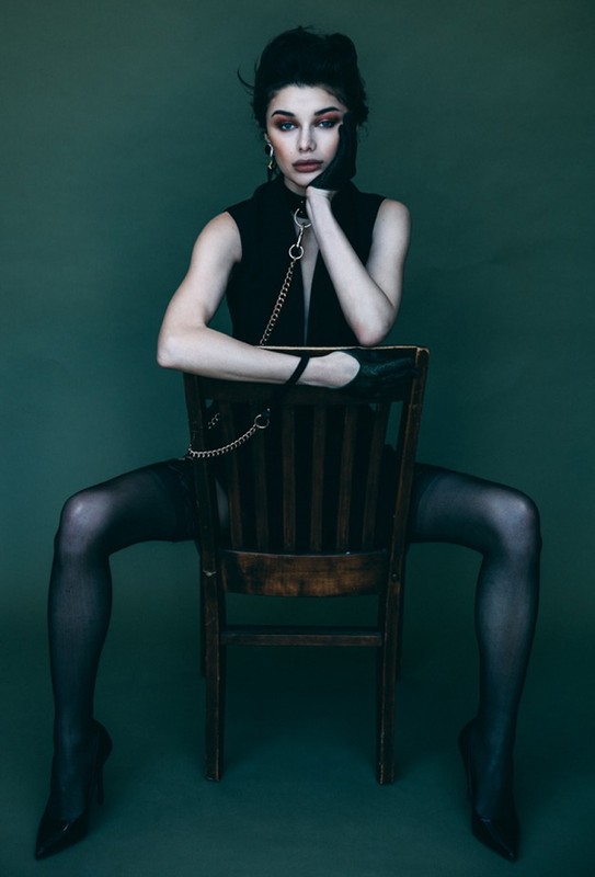 Brunette woman in a sleeveless top sits on a wooden chair with a green backdrop, wearing black stockings and heels.