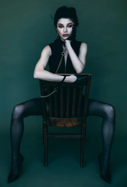 Brunette woman in a sleeveless top sits on a wooden chair with a green backdrop, wearing black stockings and heels.