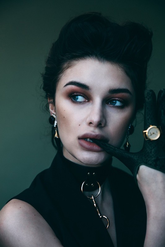 Close-up portrait of a young woman with dark hair, wearing a black choker and vest, with dramatic makeup and a hand near her lips.