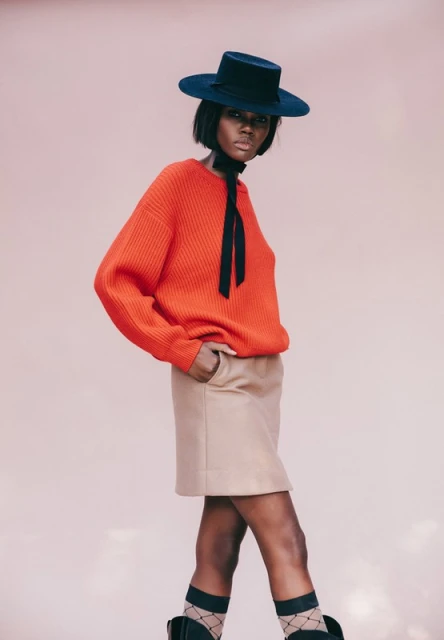 Fashion shot of a Black woman in a studio wearing a red knit sweater, tan skirt, black hat, and patterned socks against a pink backdrop.