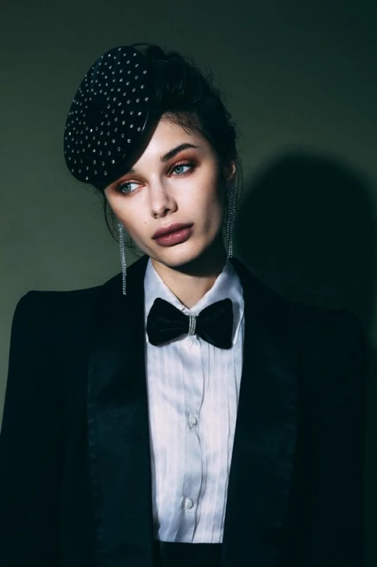 Elegant woman in a black tuxedo and beret, looking off to the side with a serene expression against a dark green background.
