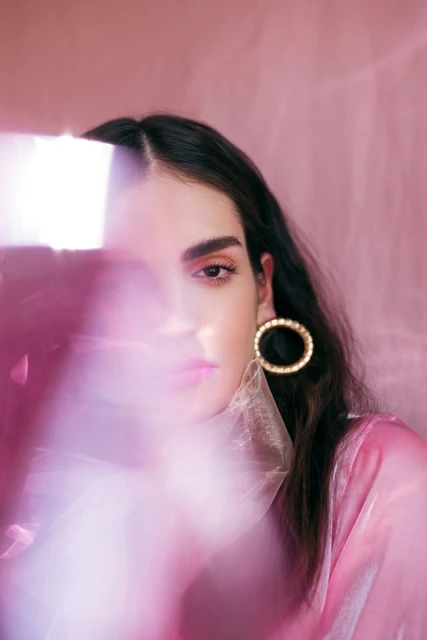 Close-up portrait of a young woman with dark hair and gold hoop earrings, partially obscured by a blurred pink filter in a pink studio.