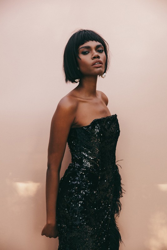 Portrait of a black woman with a dark bob haircut wearing a black sequin strapless dress against a pale background.