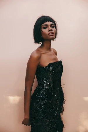 Portrait of a black woman with a dark bob haircut wearing a black sequin strapless dress against a pale background.