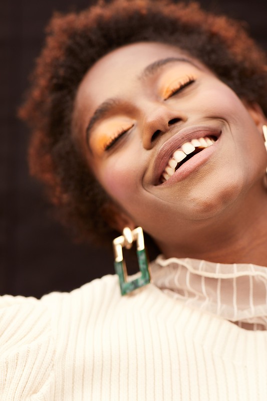 Close-up portrait of a smiling African American woman with short curly hair, gold and green earrings, and orange eyeshadow on a black background.
