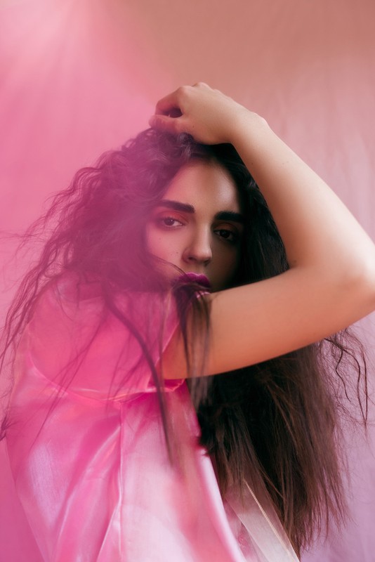 Woman with long, dark, curly hair poses against a pink backdrop, wearing a sheer pink top. She is looking at the camera.