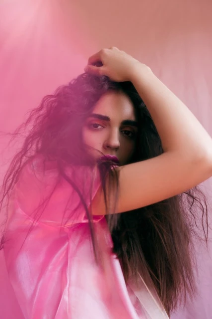 Woman with long, dark, curly hair poses against a pink backdrop, wearing a sheer pink top. She is looking at the camera.