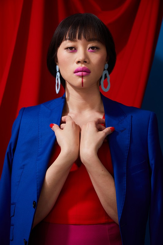 Headshot of Asian woman in blue jacket, red top, with red lipstick and graphic red stripe on her lip in front of red and blue draped backdrop.