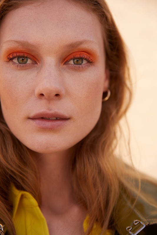 Close-up portrait of a young adult woman with red hair, wearing bright orange eyeshadow, gold hoop earrings, and a yellow shirt.