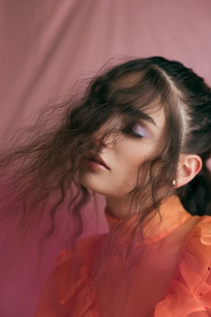 Portrait of a woman with light skin, wavy brown hair partially covering face, lilac eye shadow, and wearing a ruffled orange top.
