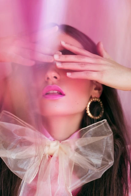 Close-up of a fashionable woman with pink lipstick covering her eyes with her hands, wearing a pink top and gold earrings.