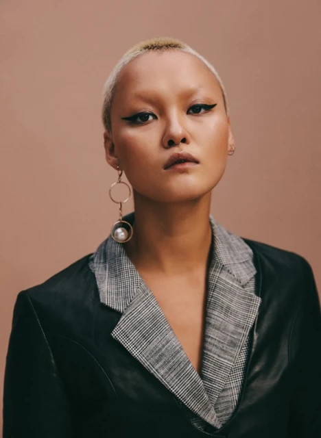 Portrait of an Asian woman with a buzzcut wearing a dark jacket with plaid lapels, set against a peach background in a studio.