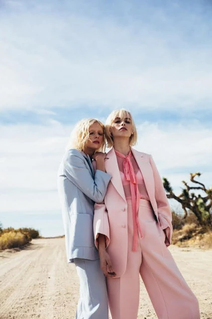 Two women pose outdoors in matching pastel suits, one in baby blue, the other in baby pink, with a desert and sky background.