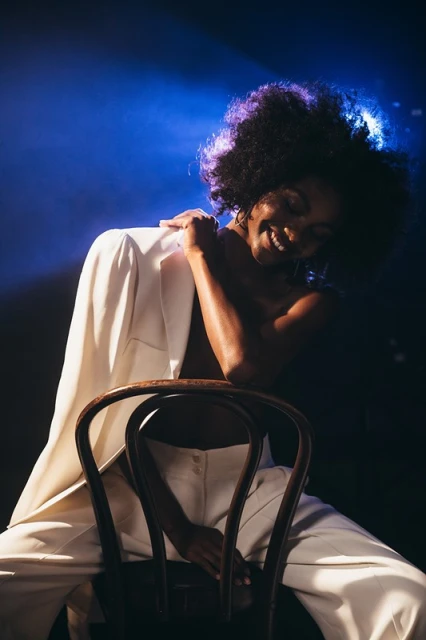 Elegant Black woman smiles sitting in white suit in dark studio with blue lighting, fashion portrait. Modern fashion, beauty concept.