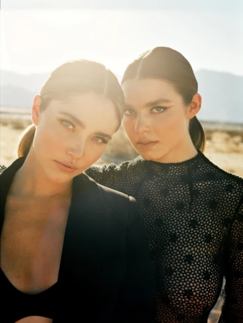 Two adult women pose closely outdoors, one in a black blazer and the other in a black mesh top against a dry, sunlit landscape.