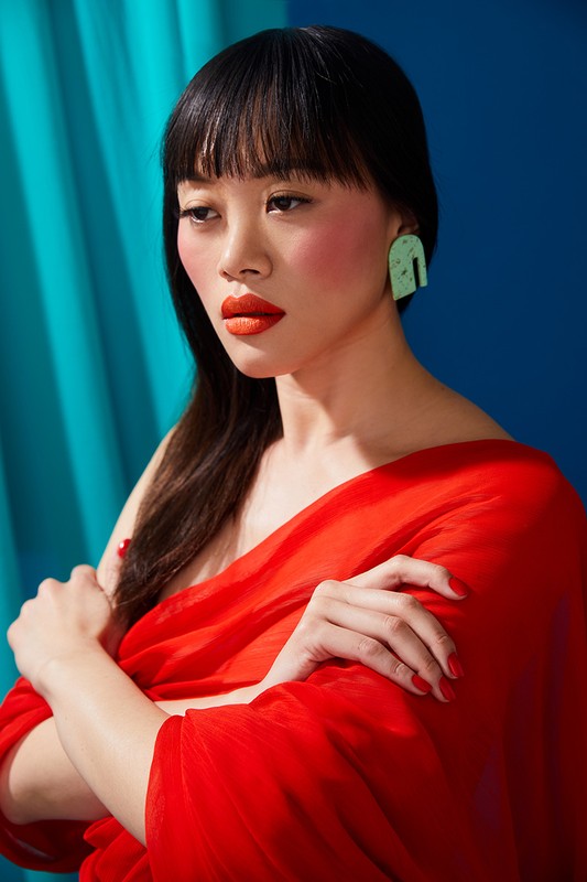 Close-up portrait of an Asian woman in a red gown against a blue backdrop, showcasing beauty and fashion elements.