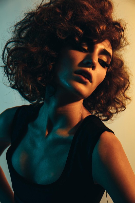 Portrait of an adult woman with curly brown hair, wearing a black top in a photo studio with cyan and yellow lighting.