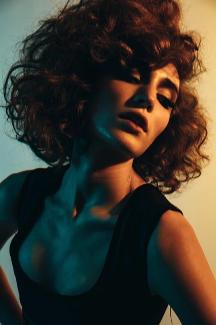 Portrait of an adult woman with curly brown hair, wearing a black top in a photo studio with cyan and yellow lighting.
