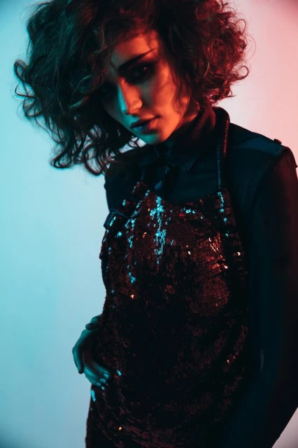 Young woman with curly dark hair and dark eye makeup poses in a sequined top with red and blue gel lighting in a studio environment.
