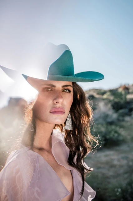 Portrait of a woman wearing a blue cowboy hat in a natural outdoor setting with light blue skies and light pink ruffled garment.