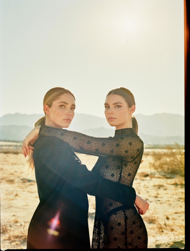 Two adult women in a desert setting, embracing in fashionable black dresses, medium shot with a soft-focus background under natural light.