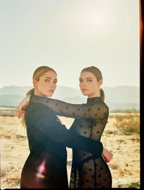 Two adult women in a desert setting, embracing in fashionable black dresses, medium shot with a soft-focus background under natural light.