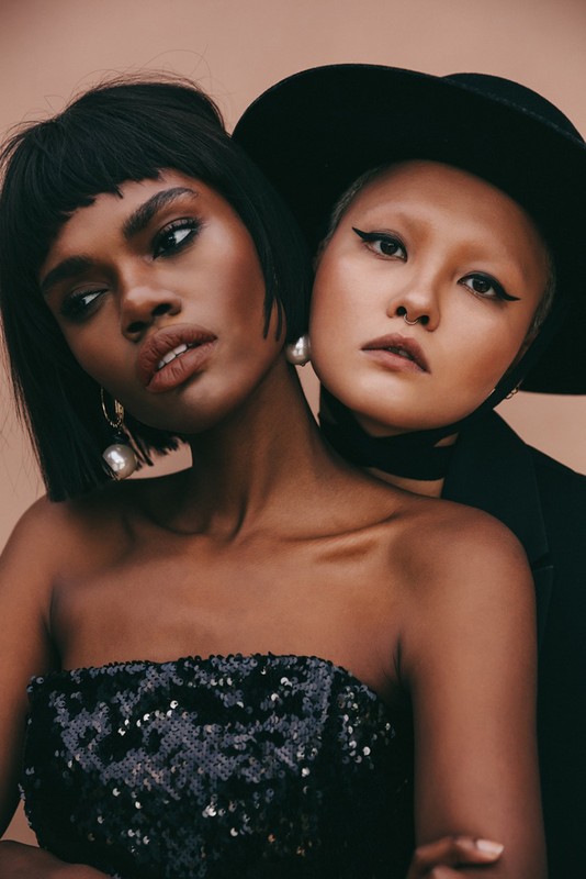 Two women pose closely together in a studio shot, one with dark hair and a sequined top, the other in a hat and jacket.