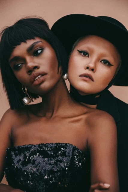 Two women pose closely together in a studio shot, one with dark hair and a sequined top, the other in a hat and jacket.