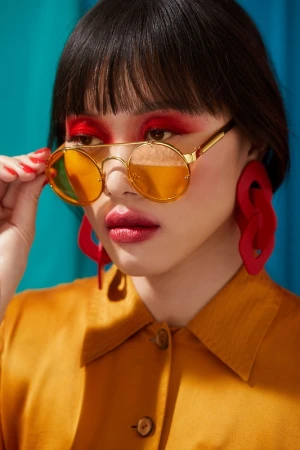 Close-up portrait of a woman wearing round yellow-tinted sunglasses, bold red eyeshadow, matching red earrings, and a mustard-yellow collared shirt.
